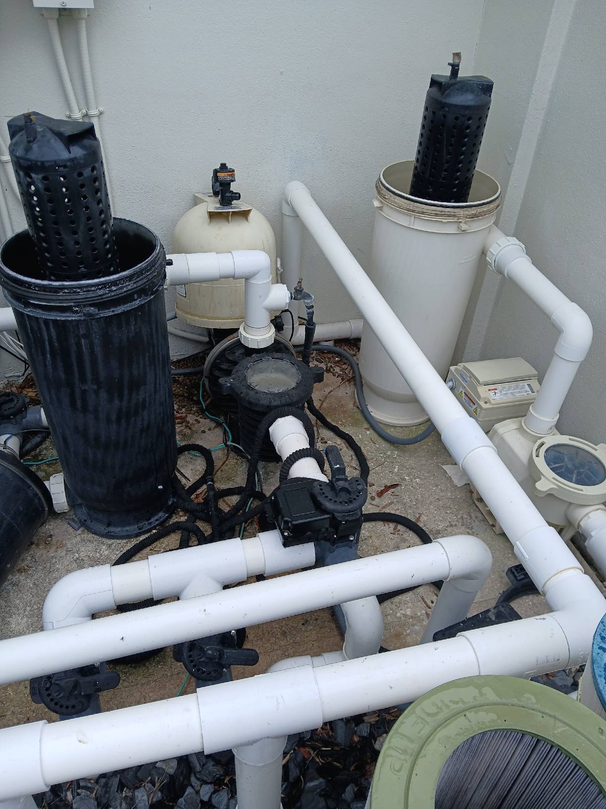 Outdoor pool equipment area showing two vertical black cylindrical filters, white PVC pipes, a round sand or cartridge filter, and a pool pump on a concrete surface.