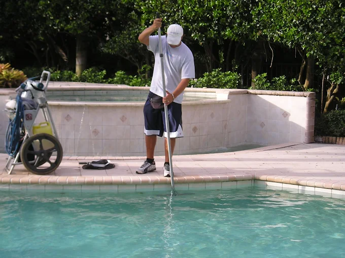 Man wearing a white cap and shirt uses a pool pole to skim or clean the pool water, beside a utility cart by the pool edge.