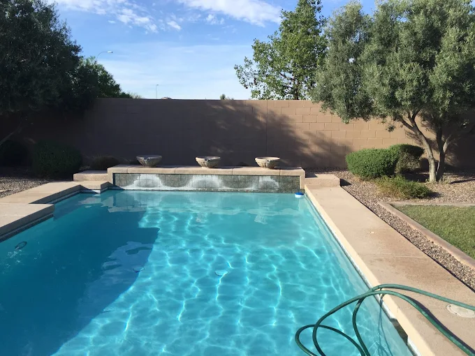 Rectangular backyard pool with a raised spa fountain and a privacy wall, clear blue water and surrounding patio.