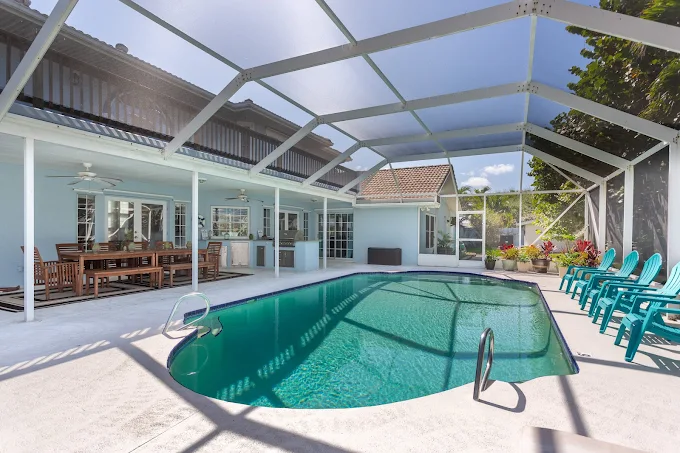Rectangular pool in a screened patio area, with a blue house in the background, a wooden dining set, and teal lounge chairs along the right side.