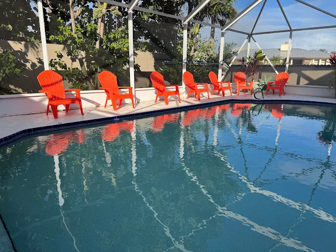 Row of bright orange Adirondack chairs along the pool deck inside a screened enclosure, with reflections in the calm water.