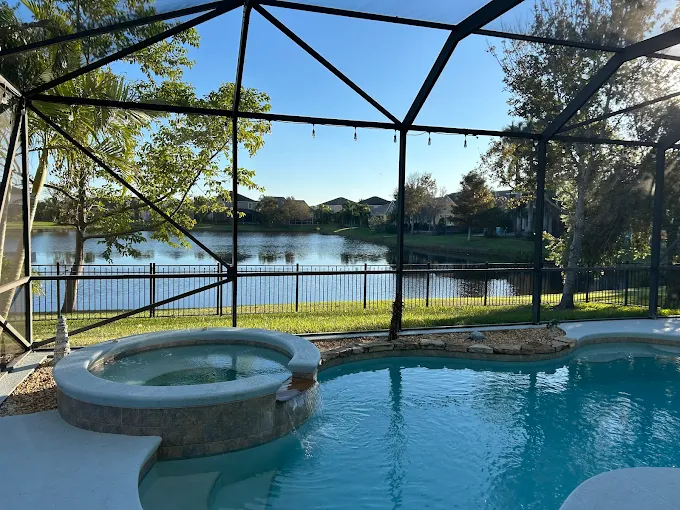 Backyard pool with a built-in spa under a screened enclosure, overlooking a lake and trees outside the fence.