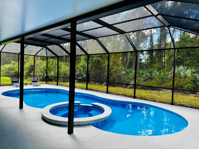Kidney-shaped blue pool with a circular attached spa, inside a screened patio enclosure with lounge chairs and greenery beyond.