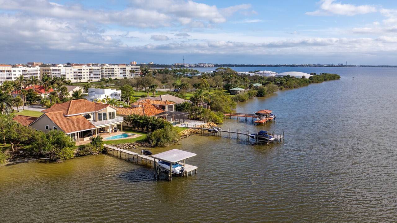 Aerial view of a waterfront residential area with red-tiled houses, palm trees, a pool, and wooden docks along calm water in a coastal town.