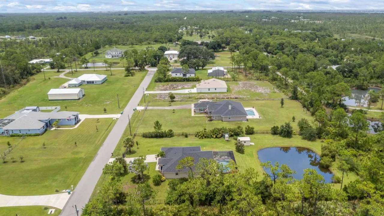 Aerial view of a suburban neighborhood with houses, green lawns, and a small pond on the right side.