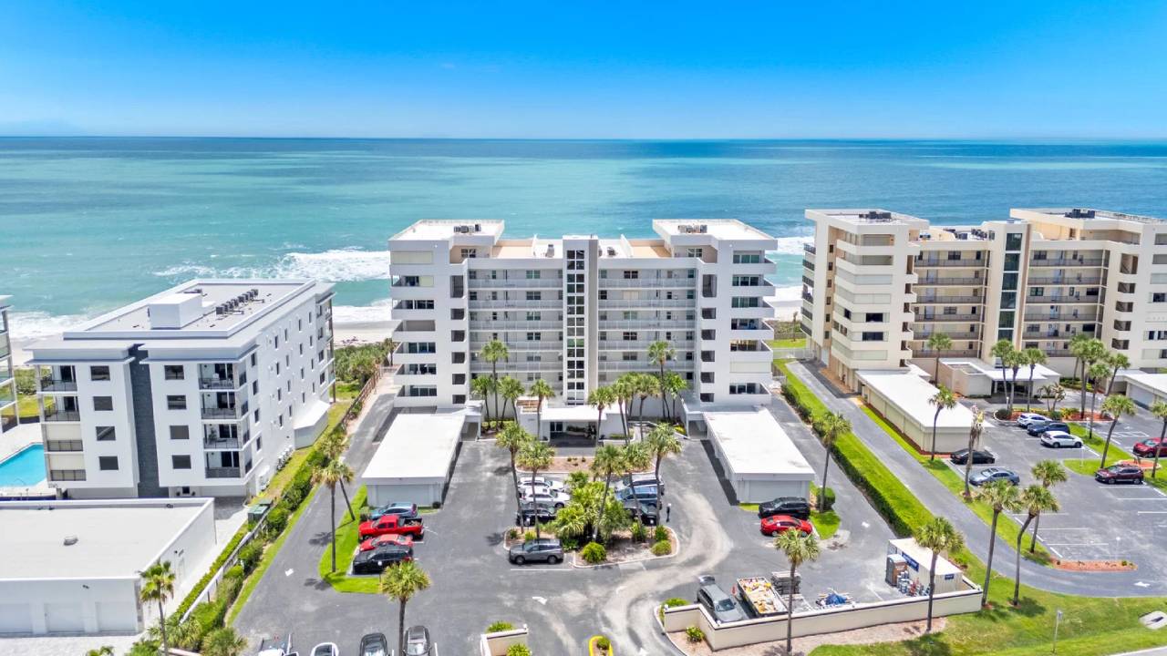 Aerial view of white beachfront condo buildings along the blue ocean, with palm trees and a parking lot in the foreground.