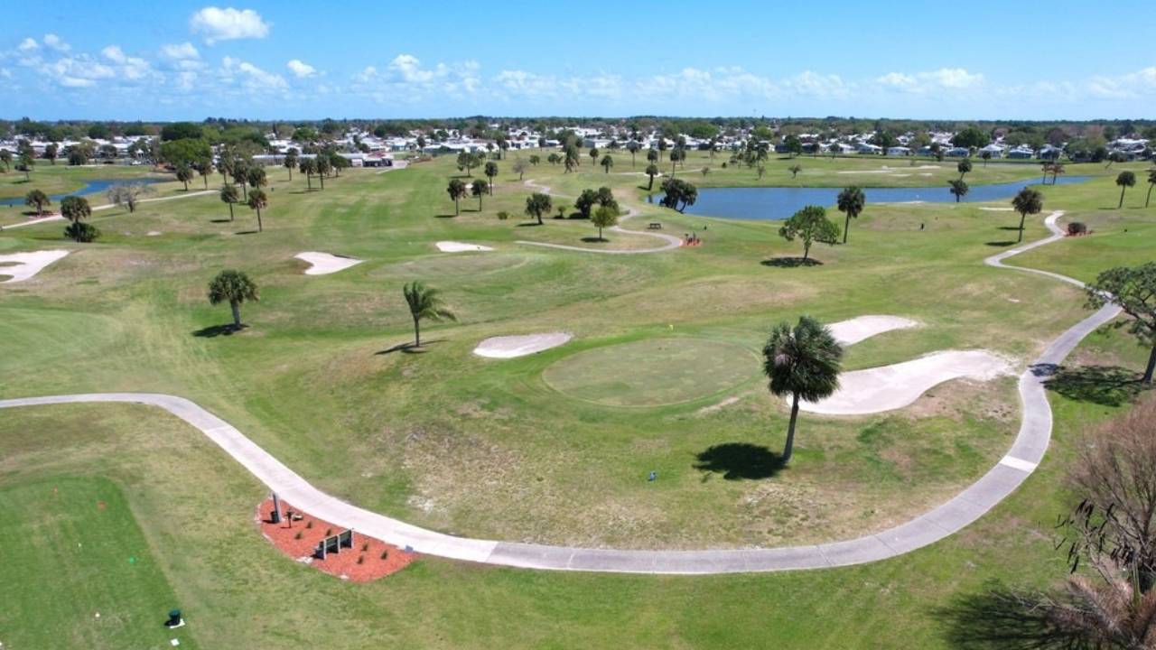 Aerial view of a golf course with greens, sand bunkers, palm trees, a winding cart path, and a small lake in the distance.