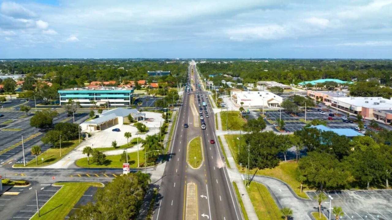 Aerial view of a long, multi-lane road flanked by shopping centers, parking lots, and green trees in a suburban area.