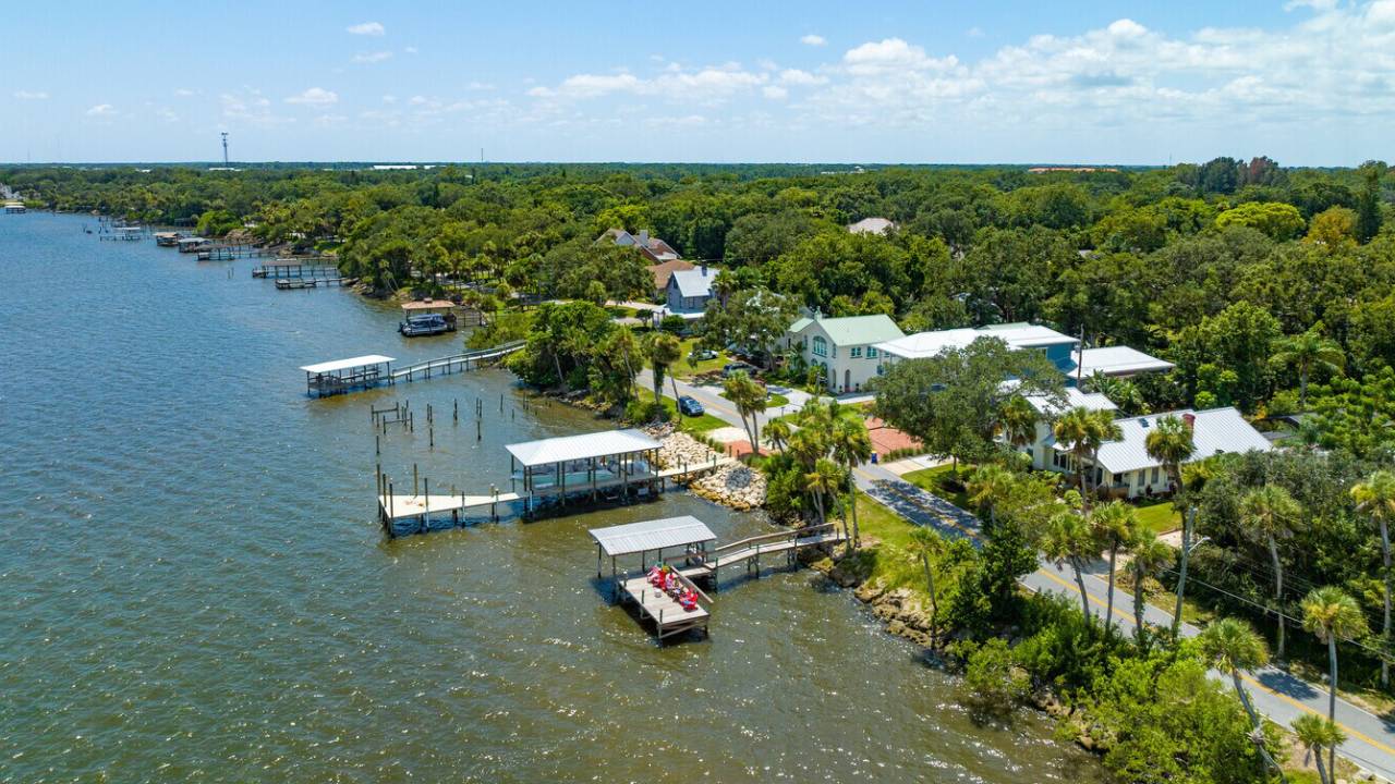 Aerial view of a waterfront residential area with houses along the shore, palm trees, and multiple boat docks along the waterway.