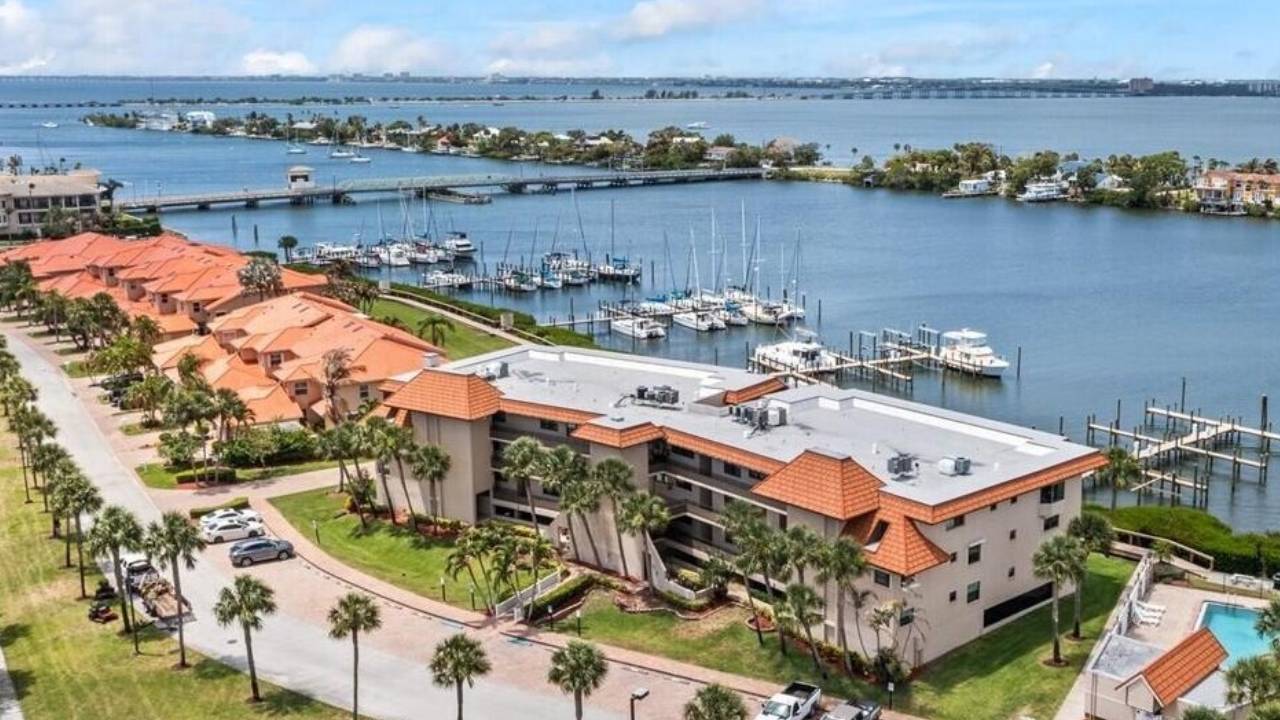 Aerial view of a waterfront residential complex with orange-roofed buildings, palm trees, and a marina with sailboats along the docks by calm blue water.