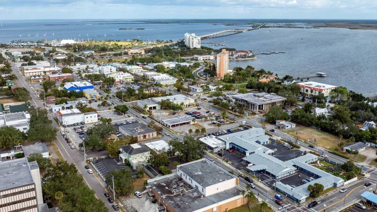 Aerial view of a coastal town with grid streets, low-rise buildings, and a bridge over water in the distance.